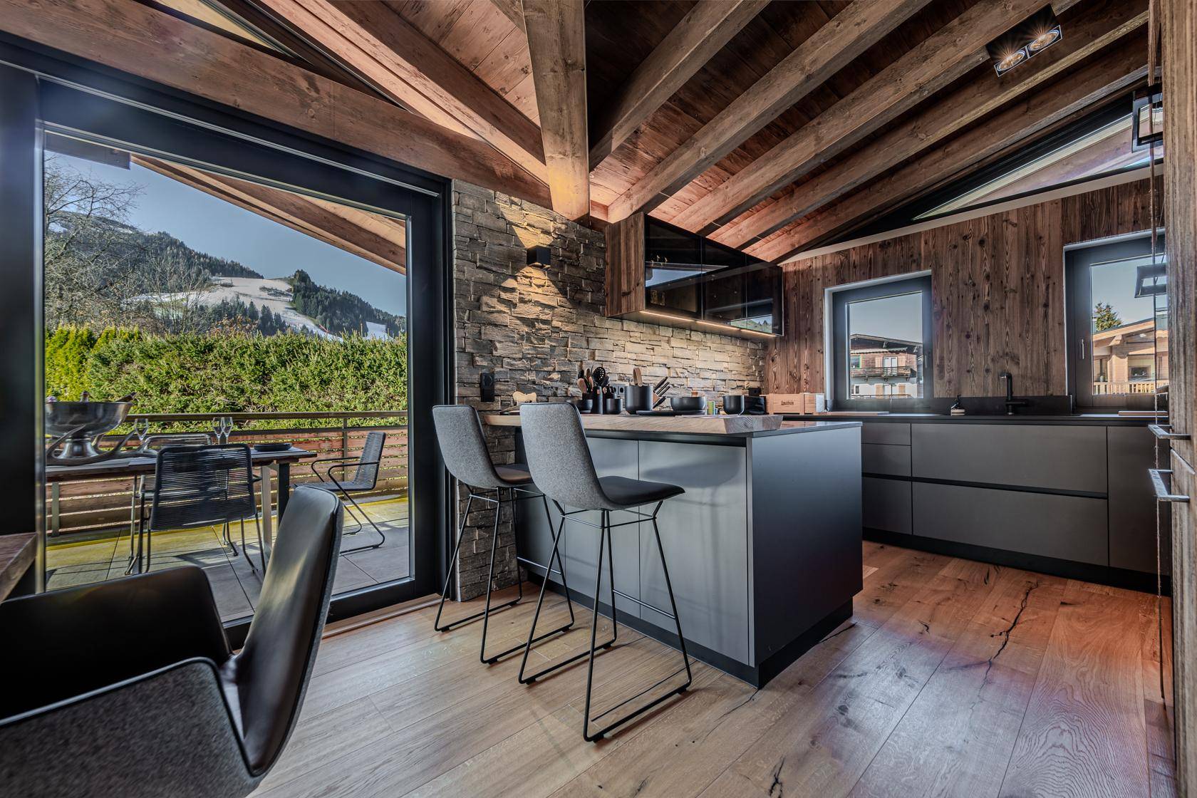 Moderne Küche mit dunklen Fronten, Steinwand und Zugang zur Terrasse mit Bergblick.