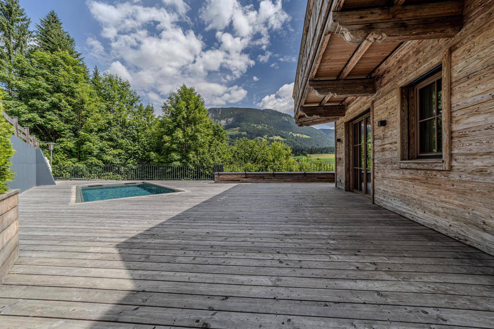 Weitläufige Holzterrasse mit Pool und Blick auf die grüne Umgebung und die Berge.