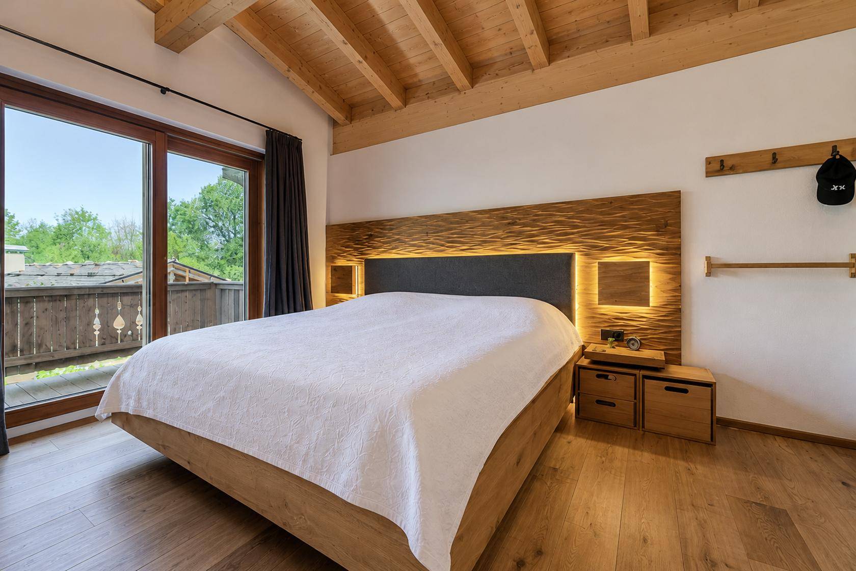 Schlafzimmer mit Holzbalkendecke, indirekter Beleuchtung und Zugang zu einem Balkon.