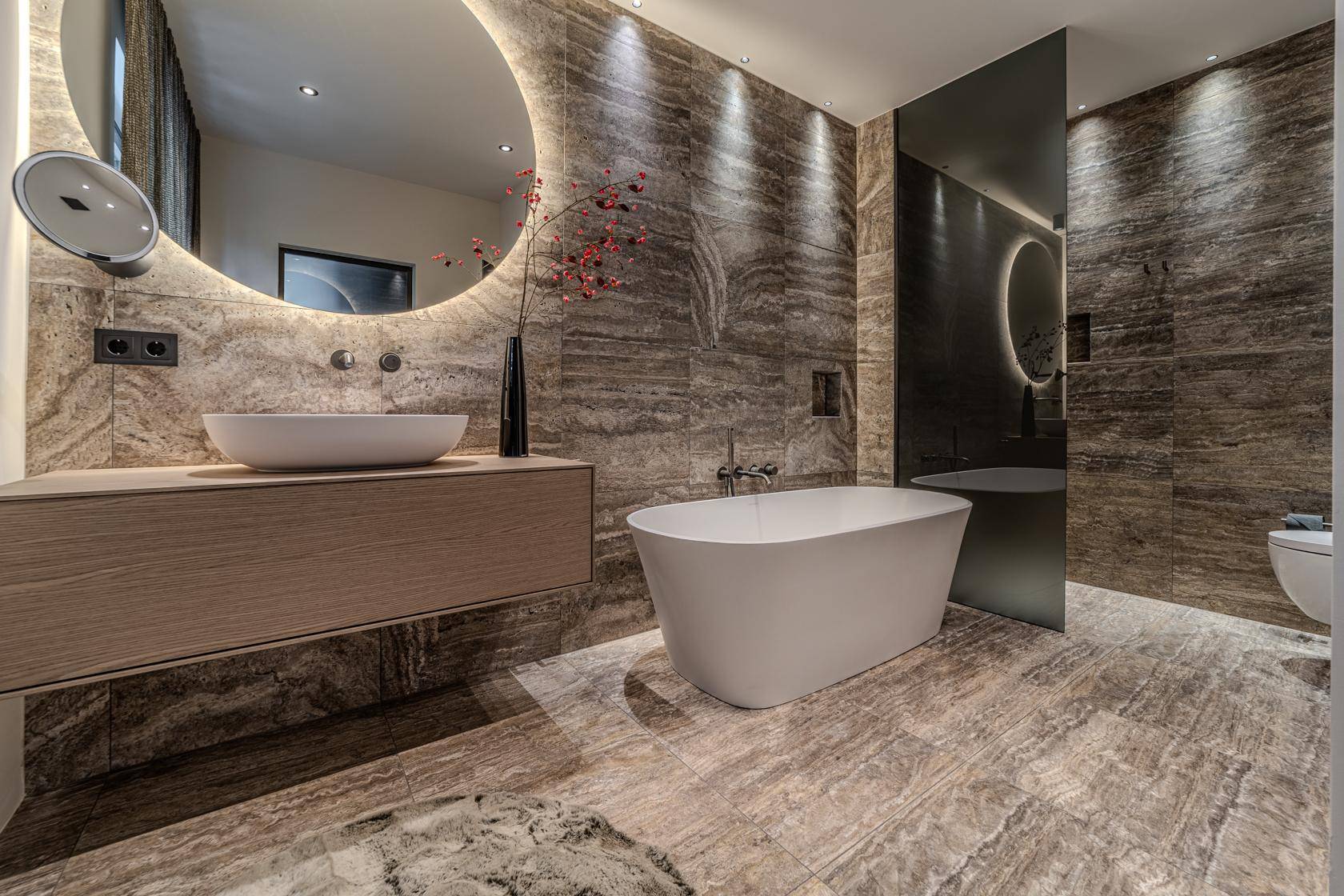 Elegantes Badezimmer mit freistehender Badewanne, großem beleuchteten Spiegel und hochwertigen Steinfliesen.