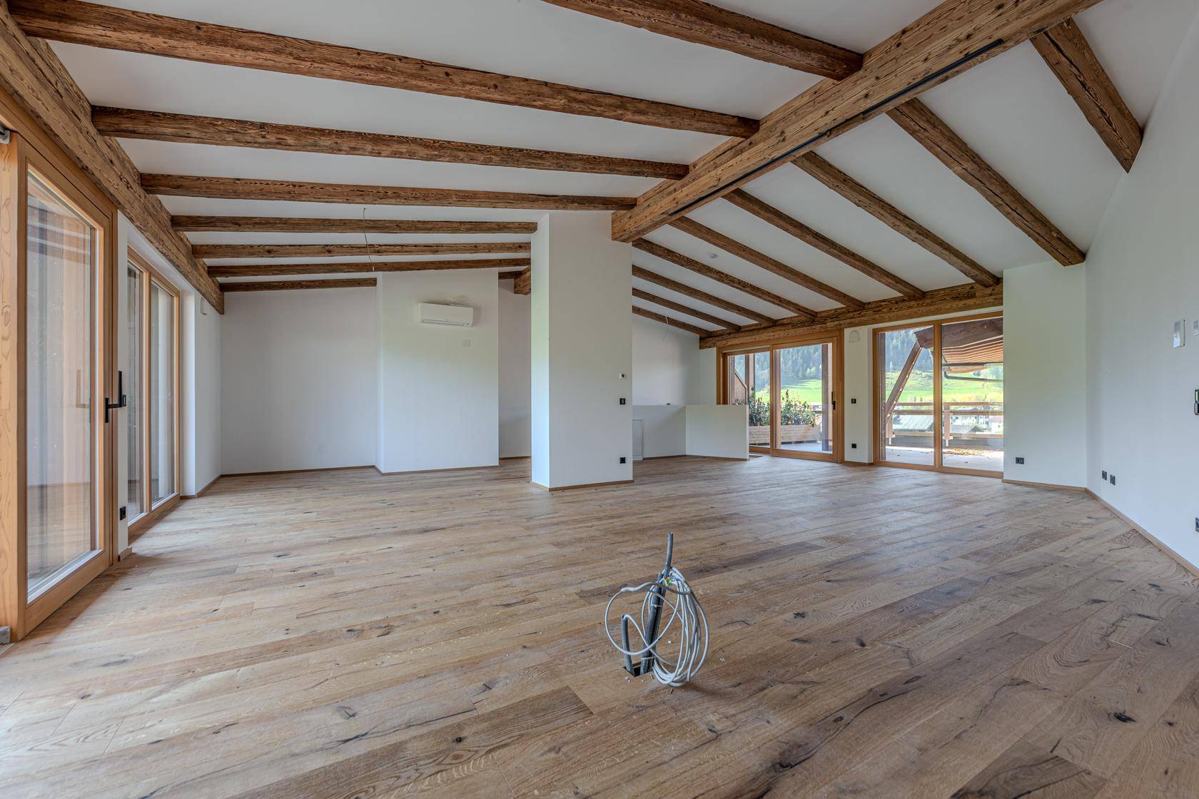 Geräumiger, heller Wohnbereich mit Holzboden, sichtbaren Deckenbalken und großen Fenstern.
