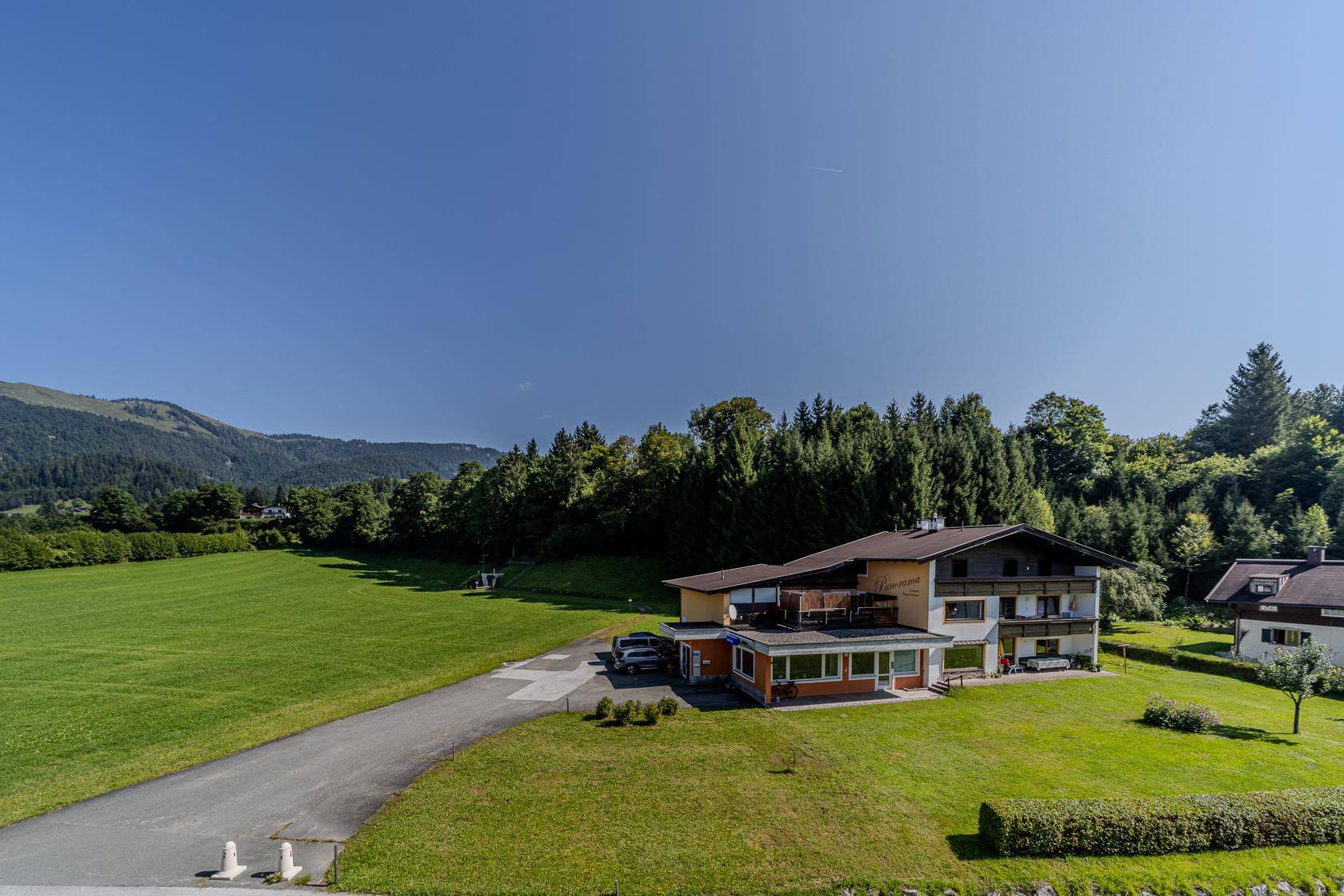 Traditionelles Gebäude in ländlicher Umgebung mit weitläufigen Grünflächen und Wald im Hintergrund.