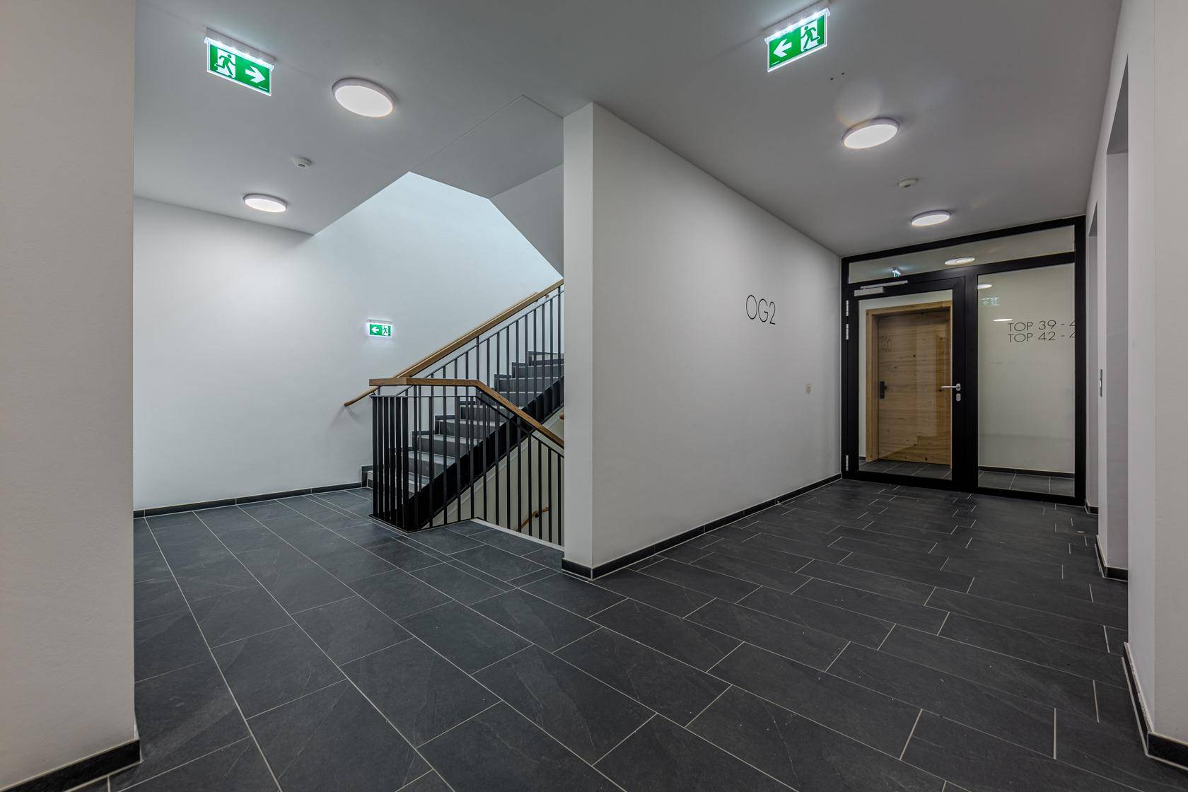 Moderner Flur mit dunklen Fliesen, weiß gestrichenen Wänden und einem Treppenhaus mit Metallgeländer.