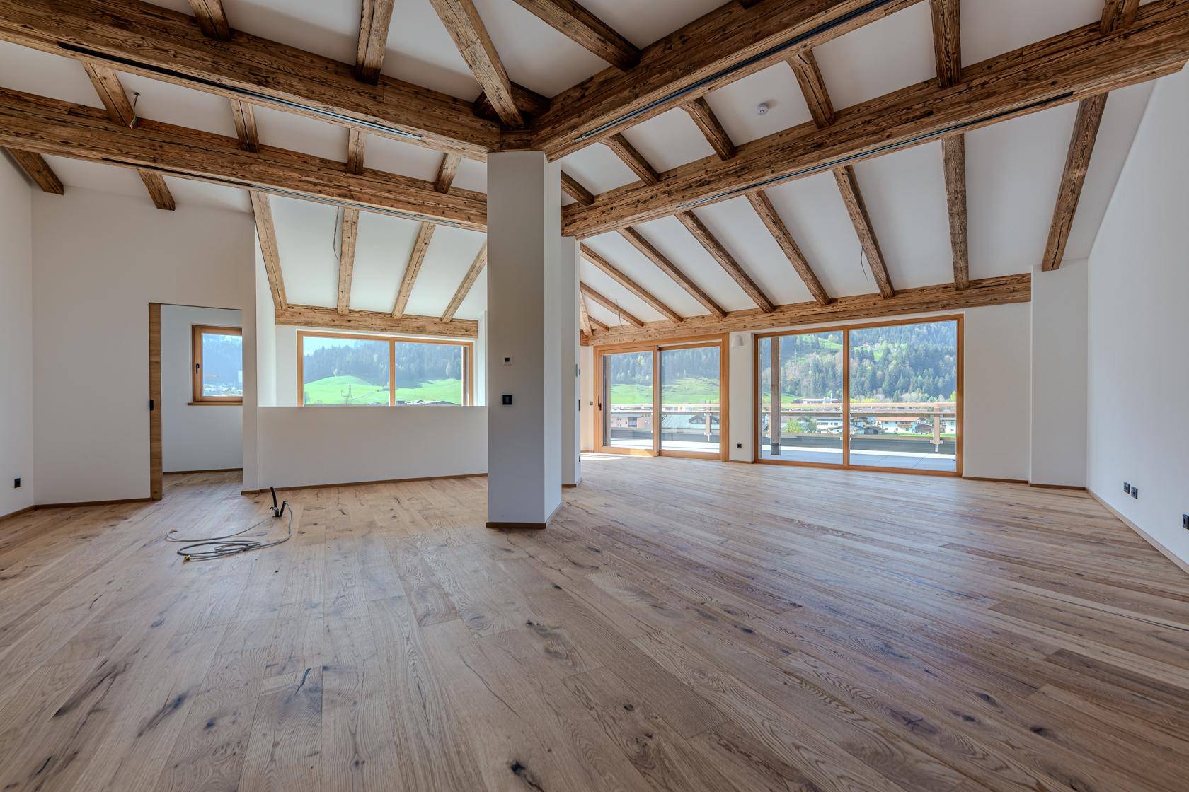 Heller, offener Raum mit Holzboden, sichtbaren Deckenbalken und großen Fenstern mit Bergblick.