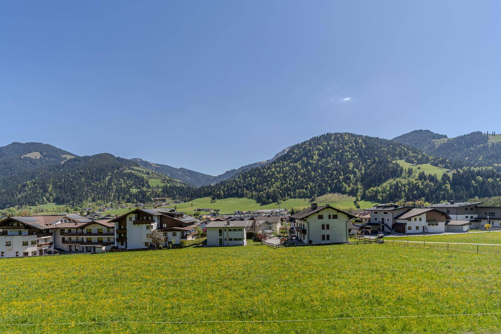 Weitläufiger Blick über eine grüne Wiese auf ein charmantes Dorf und die umliegenden Berge.