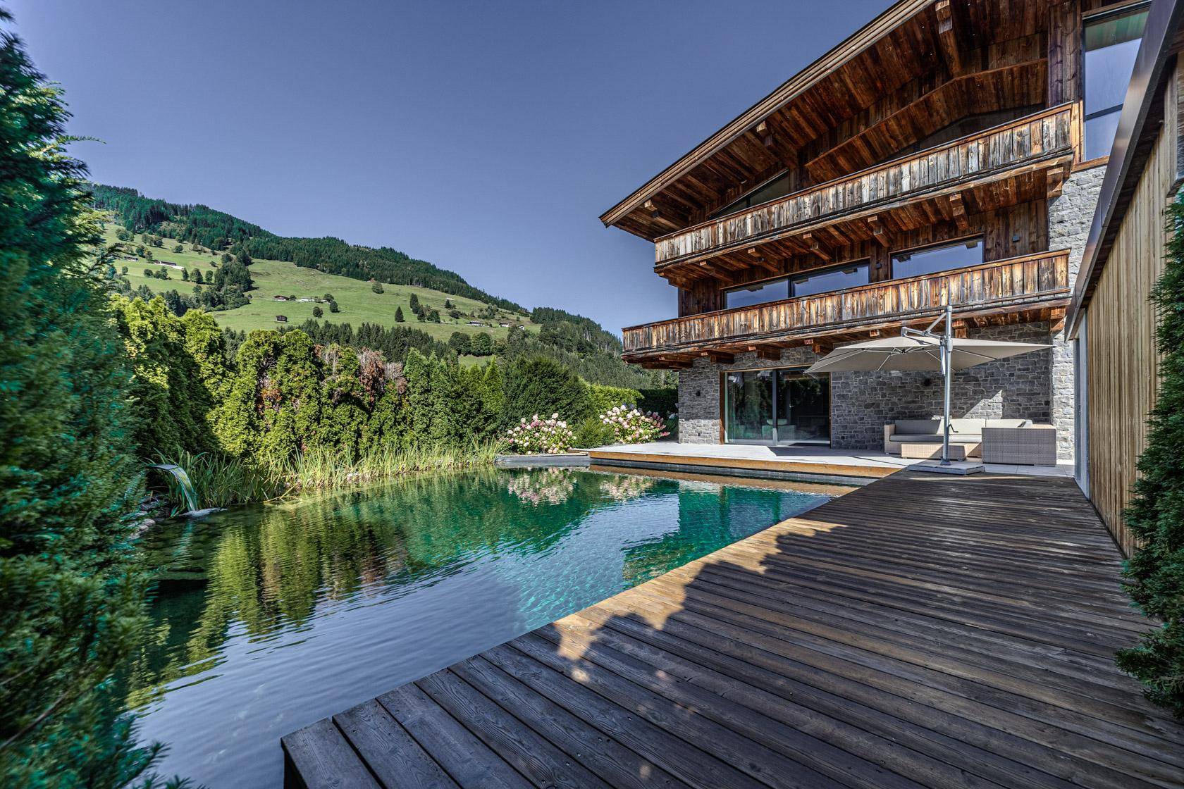 Großer Naturpool mit Holzdeck und traditioneller Holzfassade vor alpiner Bergkulisse.