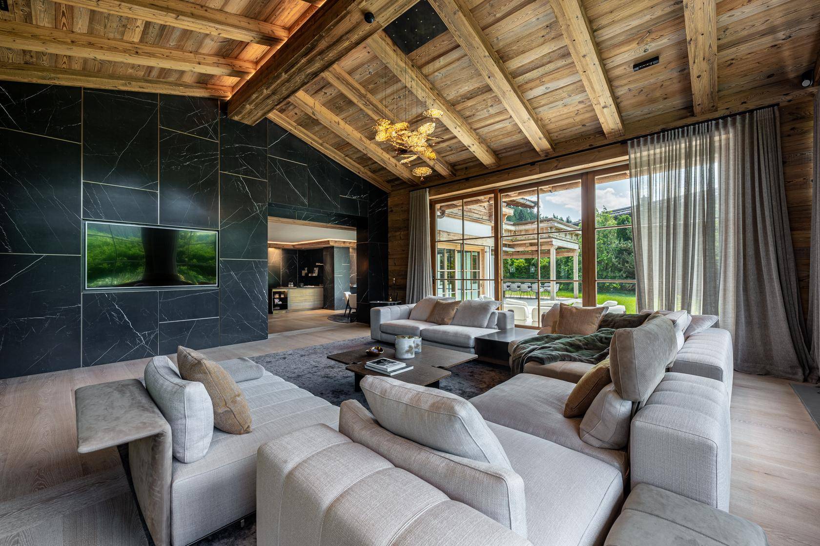 Geräumiges Wohnzimmer mit hohen Holzdecken, freiliegenden Balken und großen Fenstern mit Blick ins Grüne.
