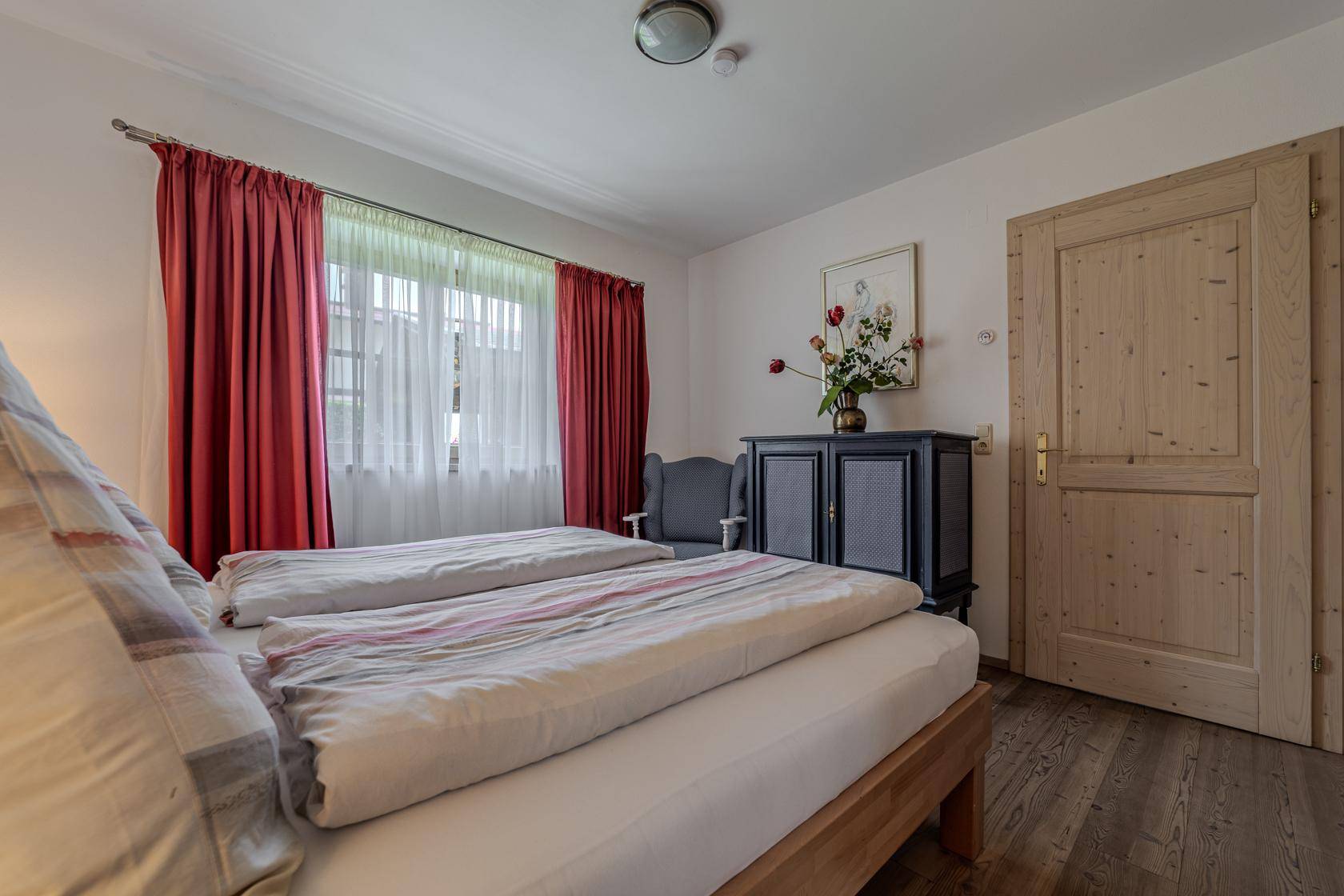 Schlafzimmer mit zwei Einzelbetten, roten Vorhängen und Holztür in hellem Ambiente.