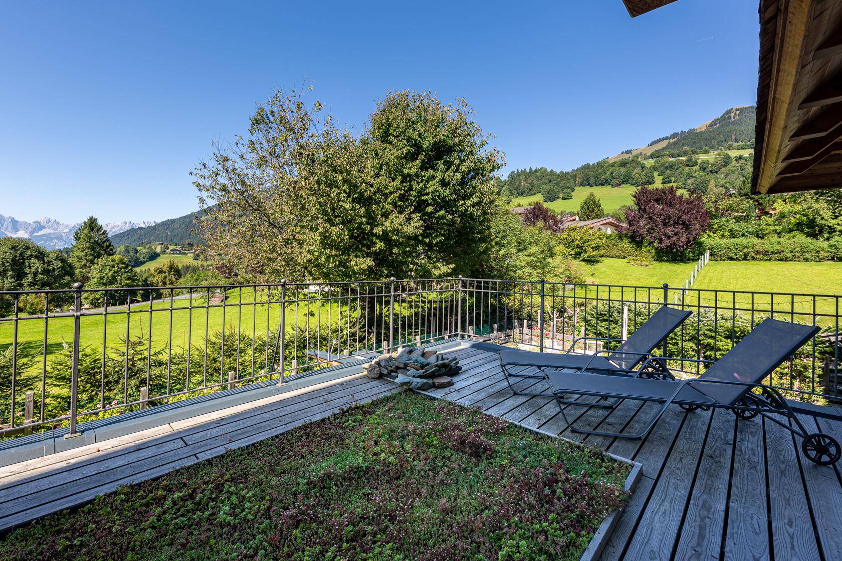 Sonnige Holzterrasse mit Liegestühlen, begrünten Flächen und weitem Blick auf die grüne Berglandschaft.