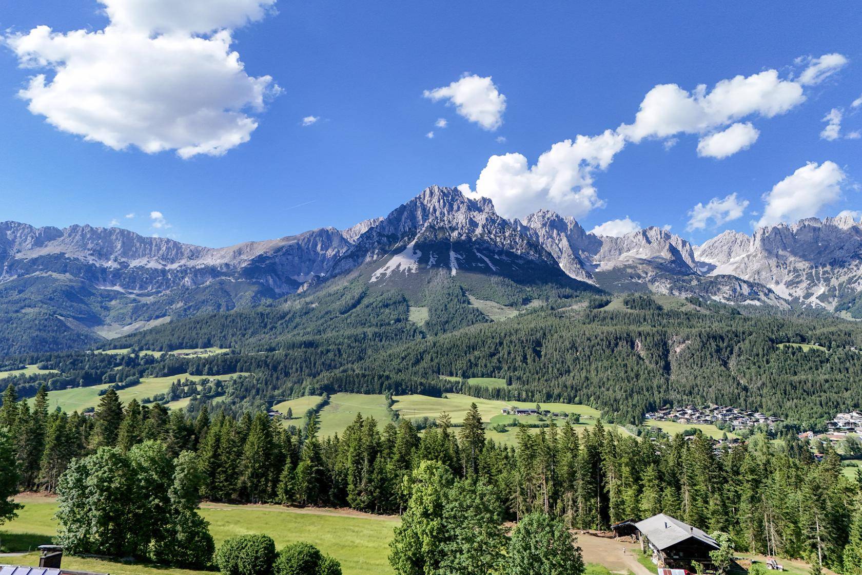 Panoramablick auf eine beeindruckende Bergkette, weite Wälder und grüne Wiesen unter blauem Himmel.