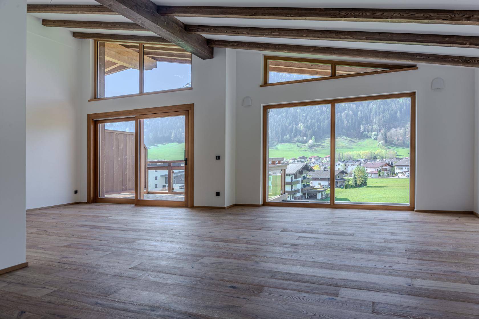 Heller Raum mit Holzboden, sichtbaren Deckenbalken und großen Fenstern mit Blick ins Grüne.
