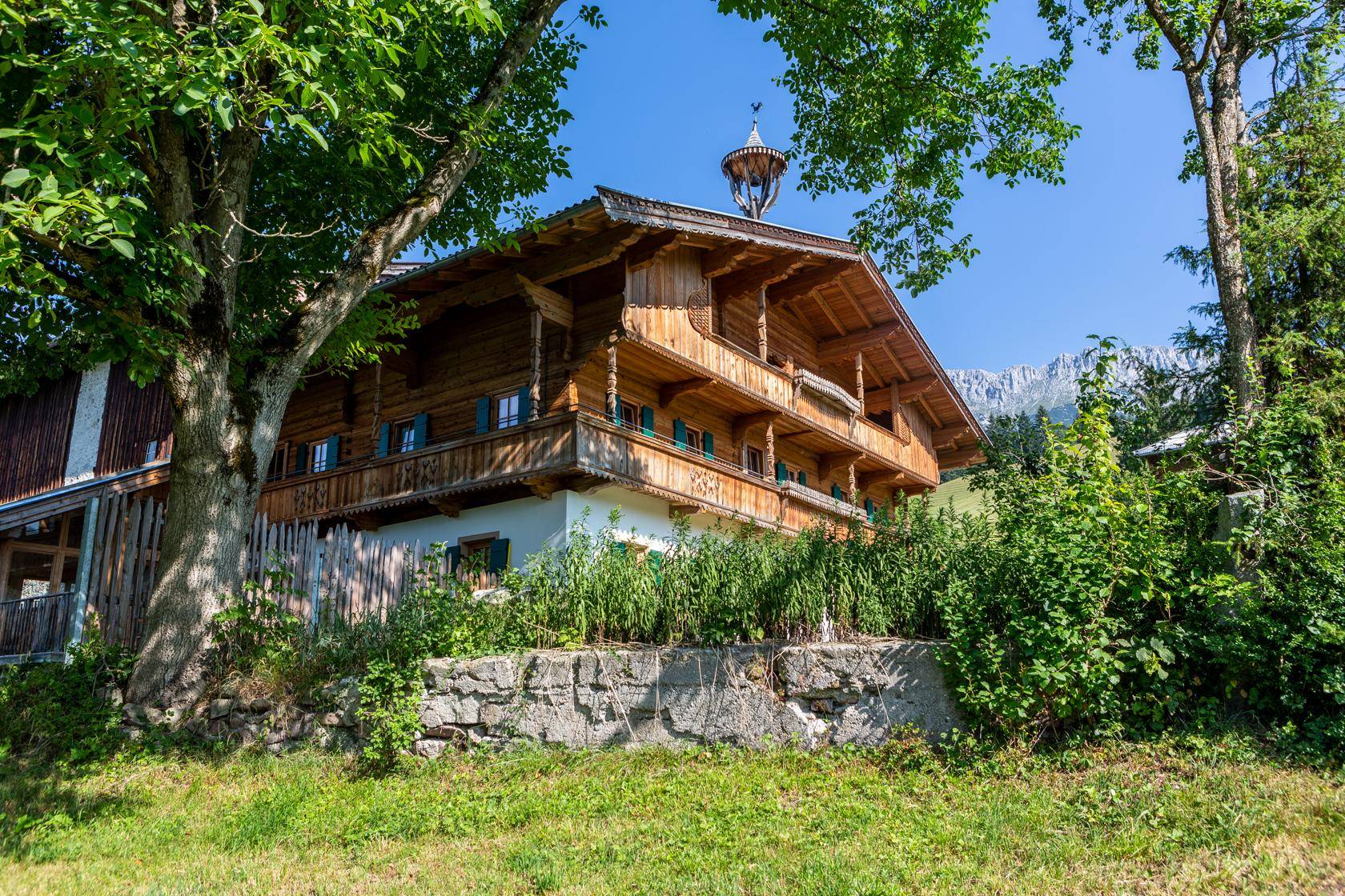 Traditionelles Holzhaus in alpiner Umgebung, umgeben von Bäumen und grüner Vegetation.