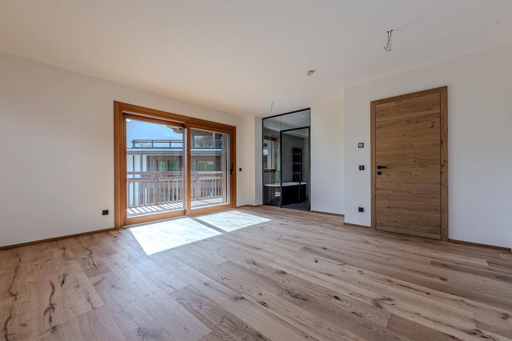 Helles Schlafzimmer mit Holzboden, Balkonzugang und Blick auf ein modernes Badezimmer.
