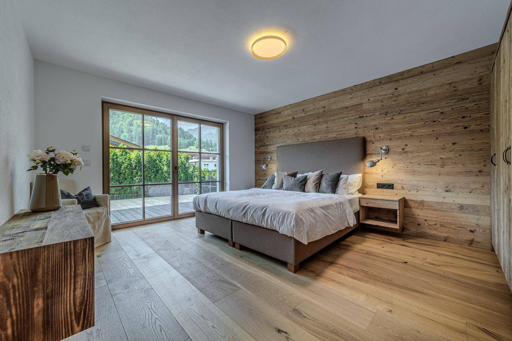 Helles Schlafzimmer mit Holzakzentwand, Holzboden und großem Schiebefenster zum Außenbereich.
