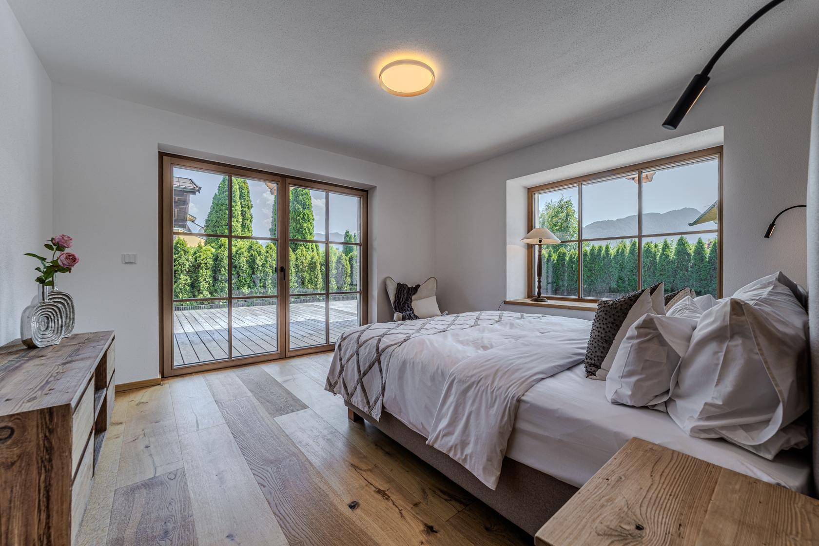 Helles Schlafzimmer mit Holzboden, großem Fenster und Zugang zu einer Terrasse mit grüner Hecke.