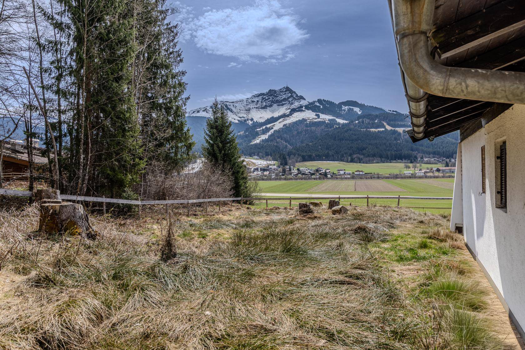 Blick auf eine weite grüne Landschaft mit schneebedeckten Bergen im Hintergrund und einem Teil des Hauses rechts.