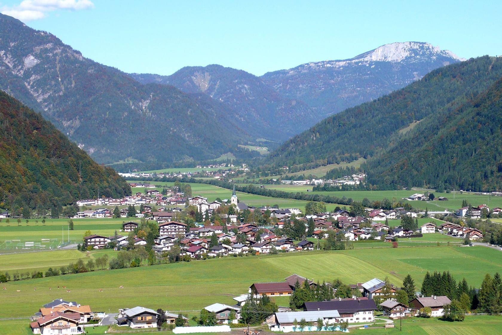 Weitläufiger Blick auf ein malerisches Tal mit Dörfern und bewaldeten Bergen unter blauem Himmel.