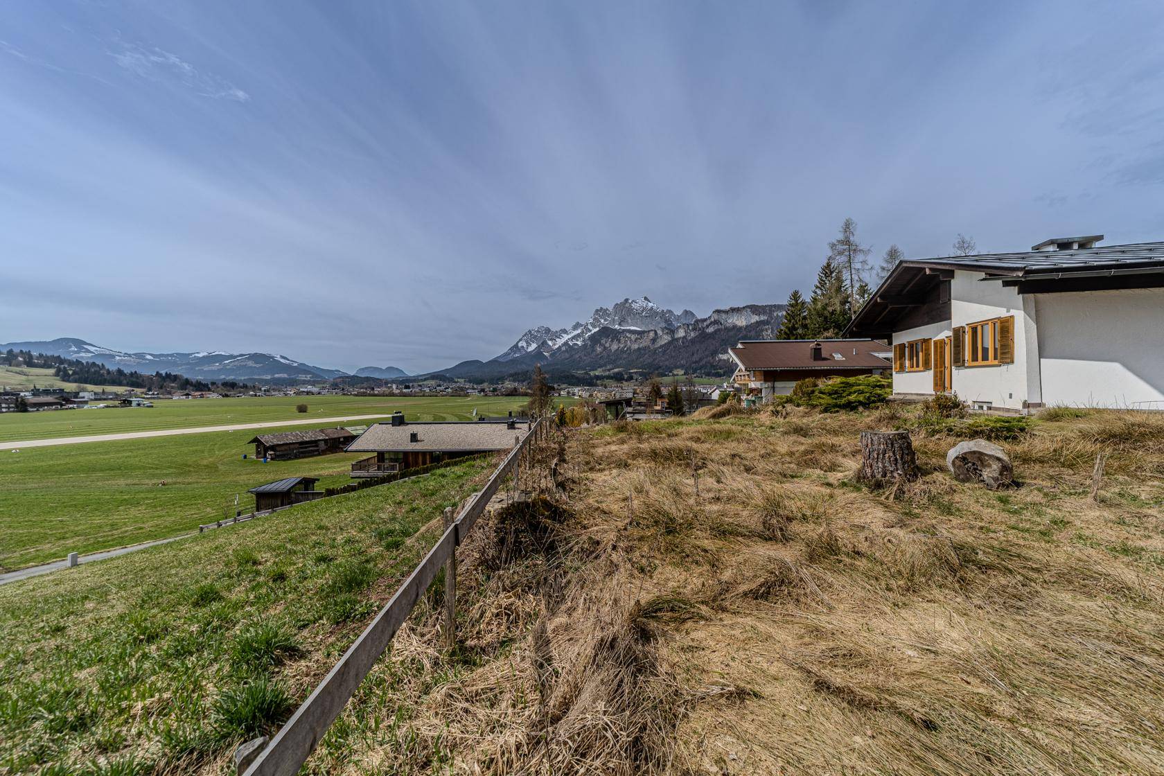 Weitläufige grüne Landschaft mit Blick auf schneebedeckte Berge und umliegende Häuser unter blauem Himmel.
