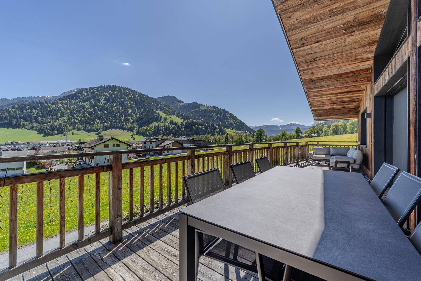 Große Holzterrasse mit Esstisch und Lounge-Bereich, umgeben von Bergen und grüner Landschaft.