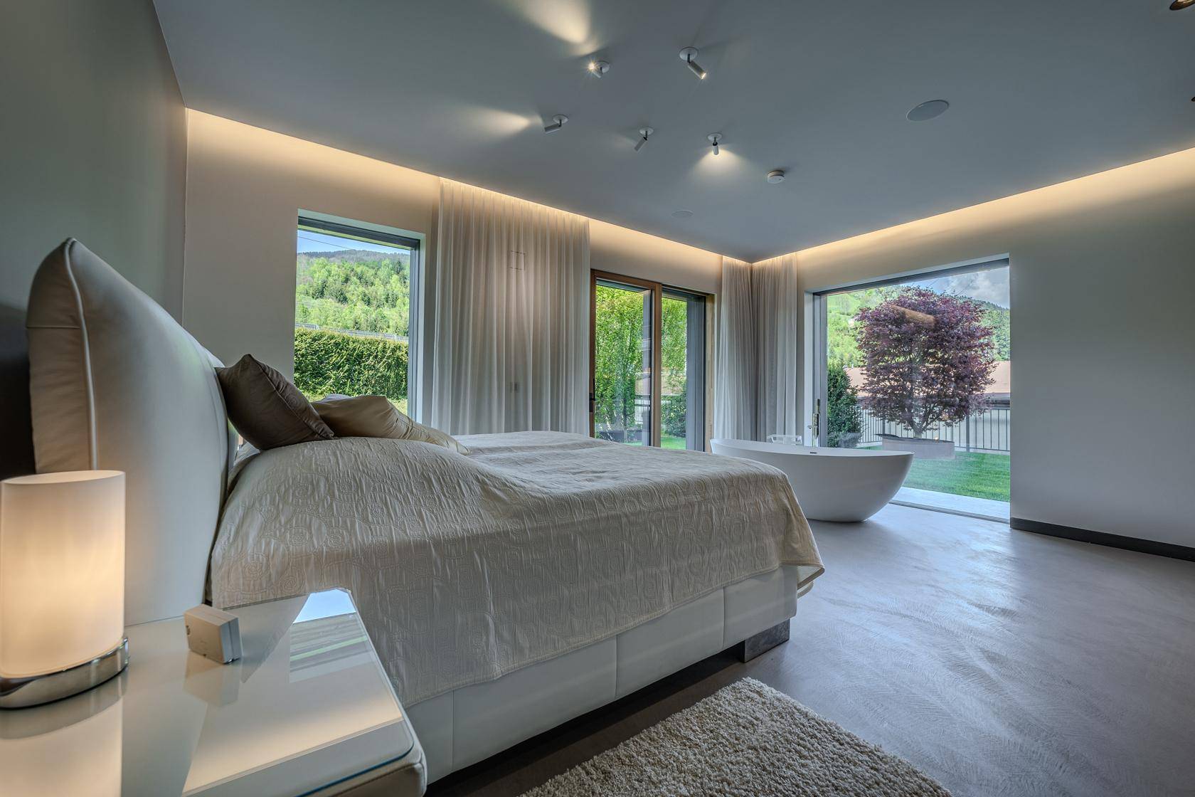 Großes Schlafzimmer mit Doppelbett, freistehender Badewanne und Panoramablick ins Grüne.