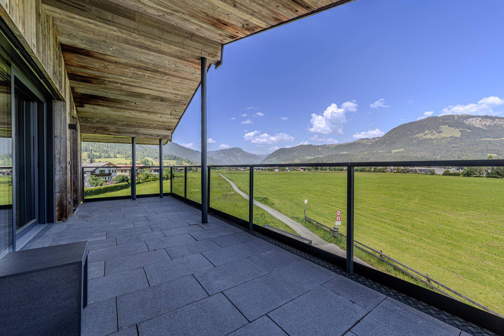 Weitläufige Terrasse mit Glasgeländer und Panoramablick auf die umliegende Berglandschaft und grüne Felder.