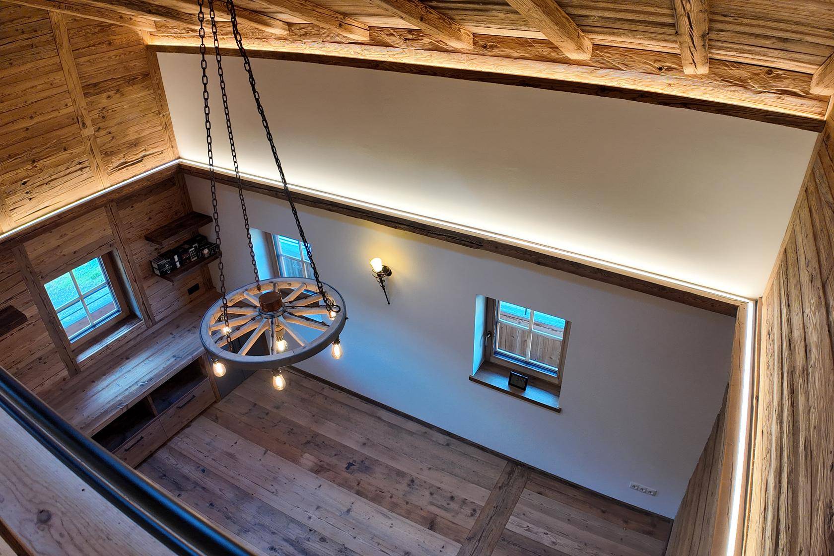 Blick von oben auf das lichtdurchflutete Wohnzimmer mit hohen Decken und rustikalem Wagenrad-Kronleuchter.