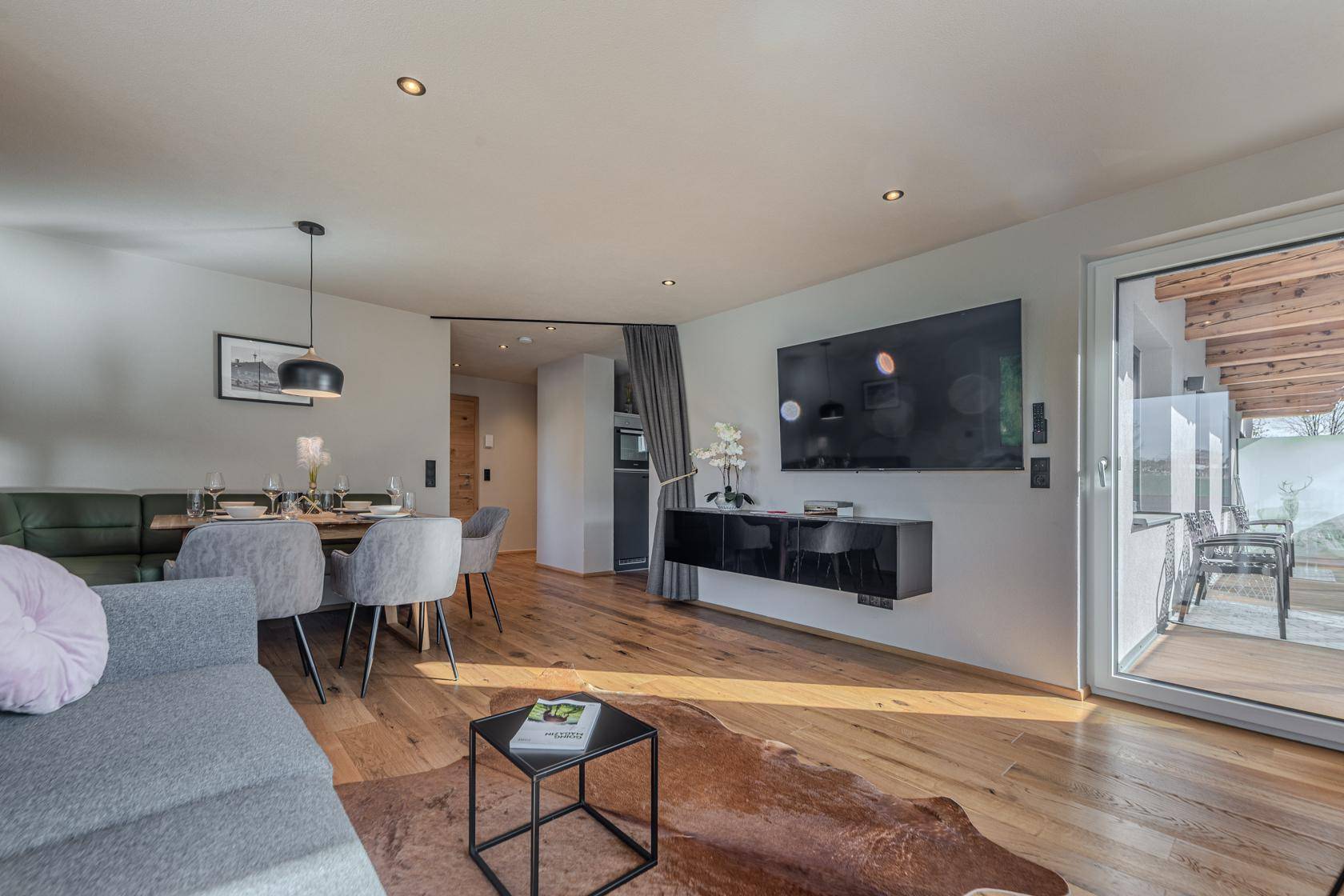 Heller Wohn-Essbereich mit Holzboden, Wandfernseher und direktem Zugang zum Balkon.