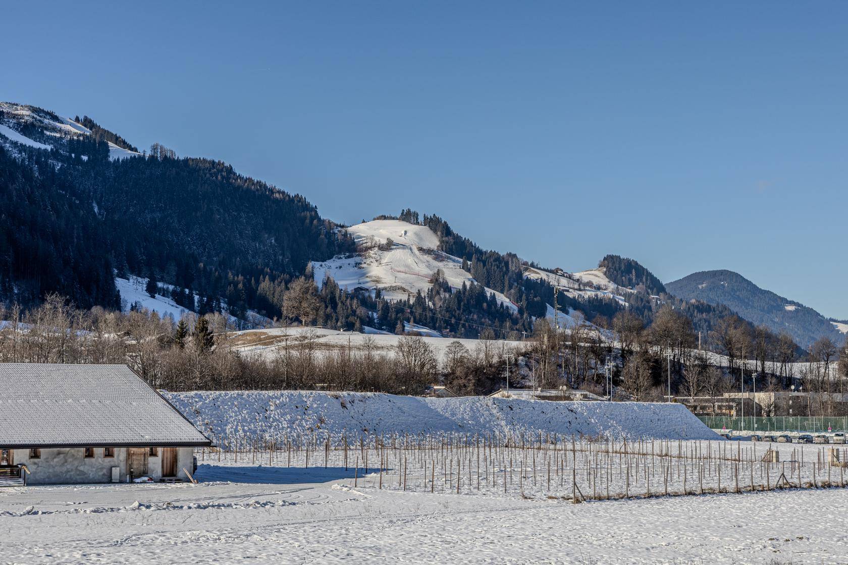 Weitläufige Winterlandschaft mit schneebedeckten Bergen und einem Bauernhof im Vordergrund.