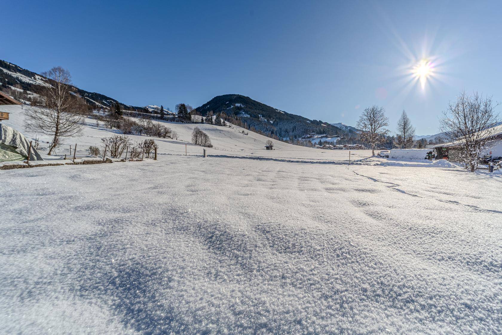 Weite, sonnige Winterlandschaft mit tief verschneiten Feldern und Bergen im Hintergrund.