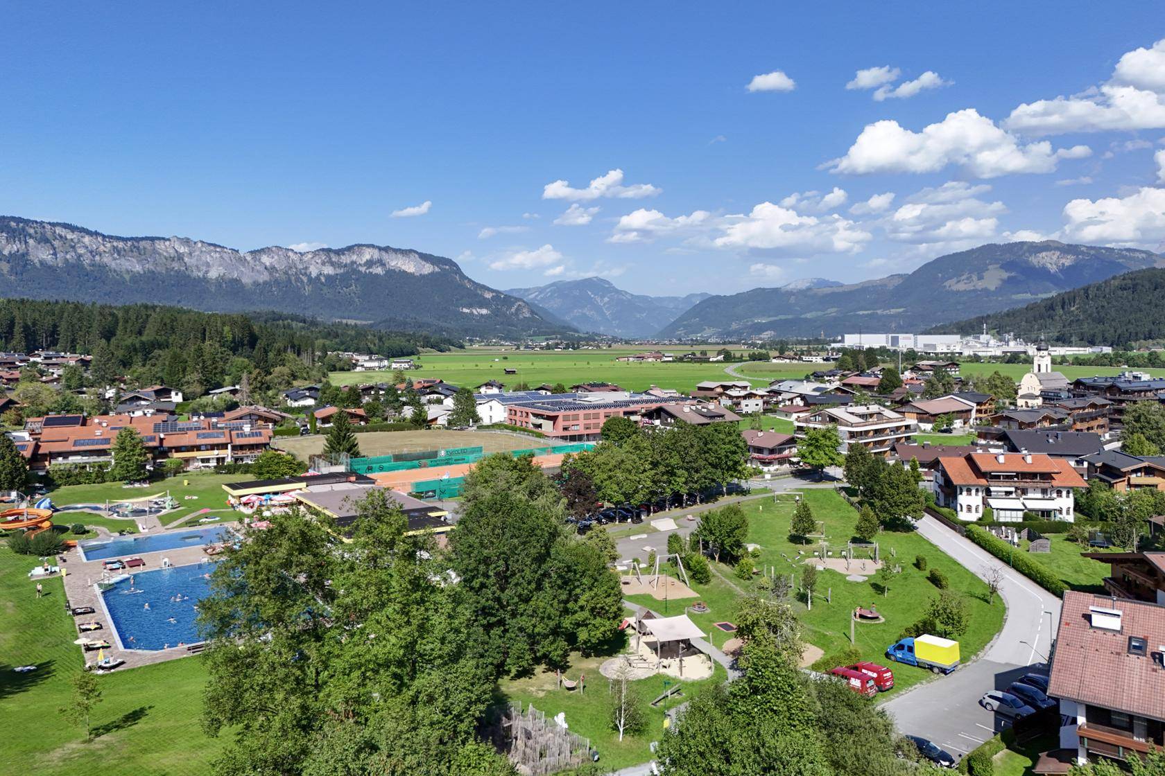 Luftaufnahme eines Dorfes mit Freibad, Tennisplatz und Spielplatz vor Bergkulisse.