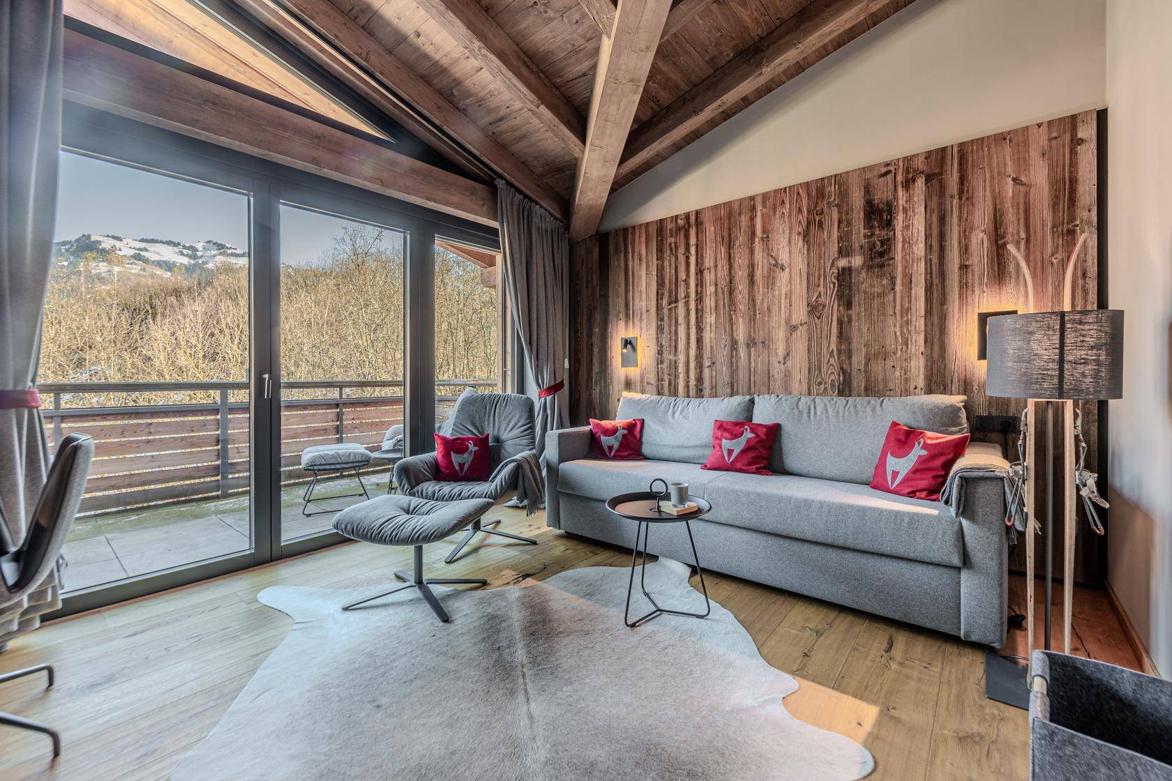 Gemütliches Wohnzimmer im Chalet-Stil mit Holzwänden, Sofa und Zugang zu einem Balkon mit Bergblick.