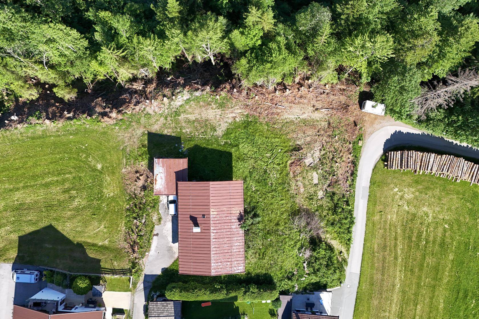 Luftaufnahme eines Hauses mit rotem Dach, umgeben von grünen Wiesen und Wald, mit Zufahrt.
