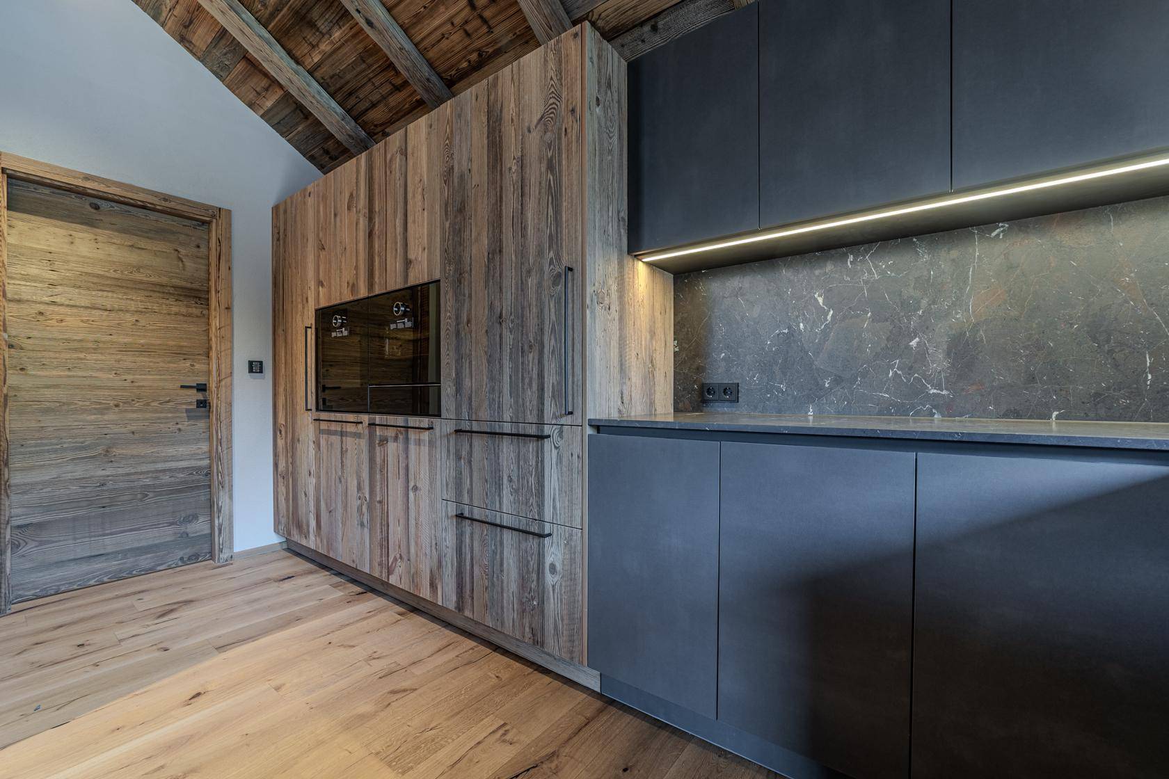 Detailansicht der modernen Küche mit integriertem Backofen, Kühlschrank und dunklen sowie hölzernen Schrankfronten.