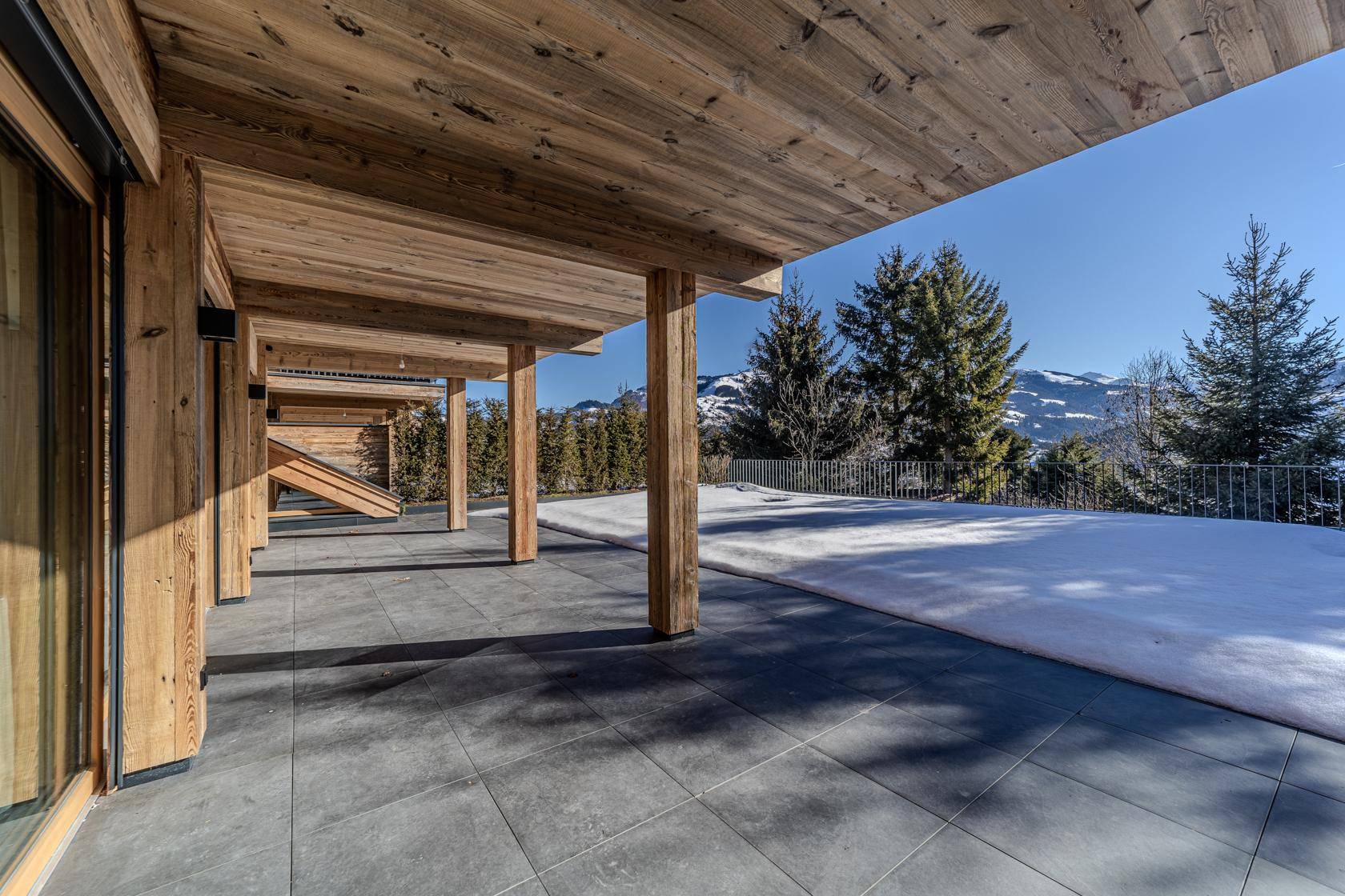 Überdachte Terrasse mit Holzbalken und Fliesenboden, umgeben von verschneiter Natur.