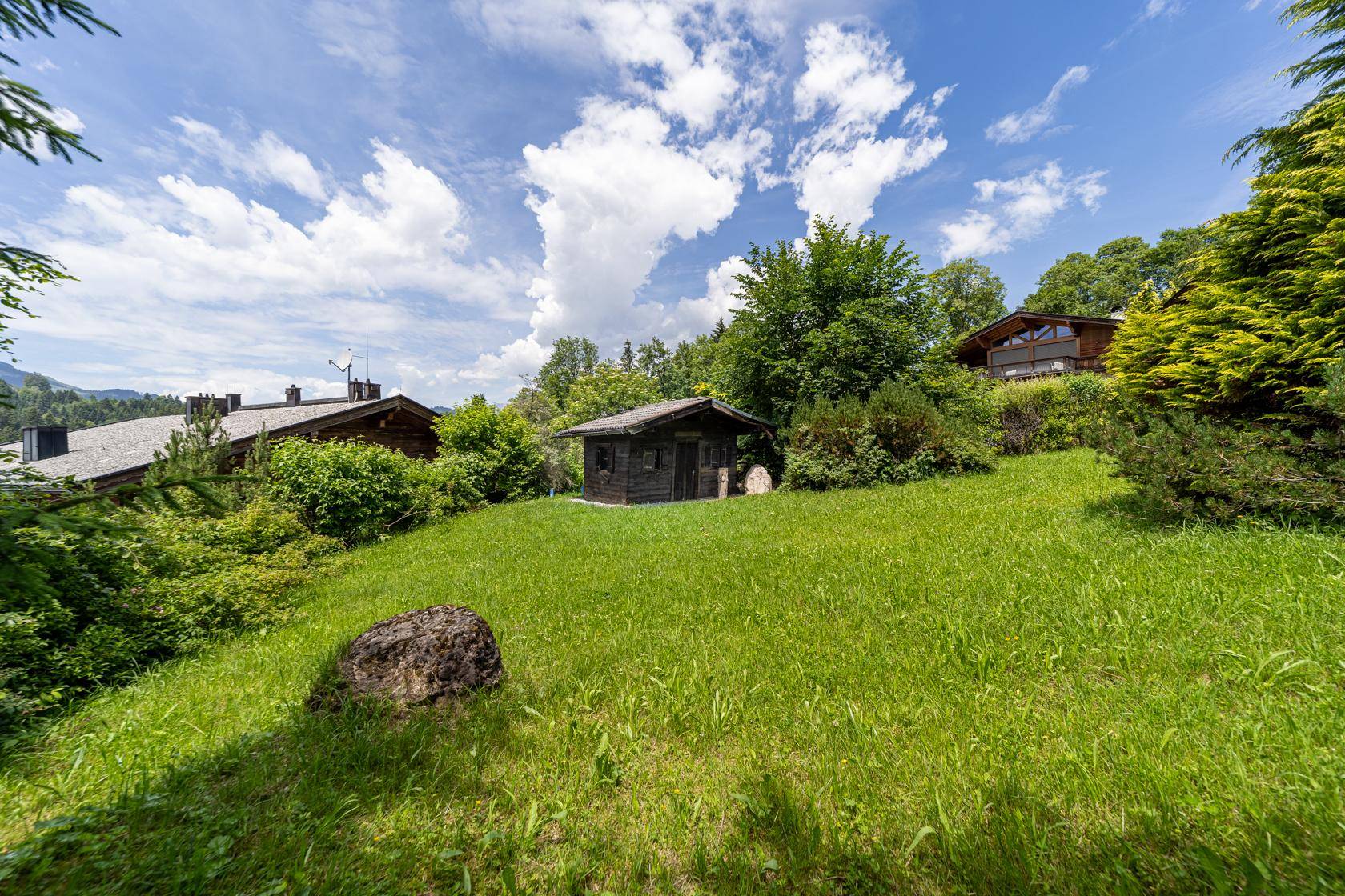 Großer Gartenbereich mit Rasenfläche, einem kleinen Holzschuppen und alpenländischer Umgebung.