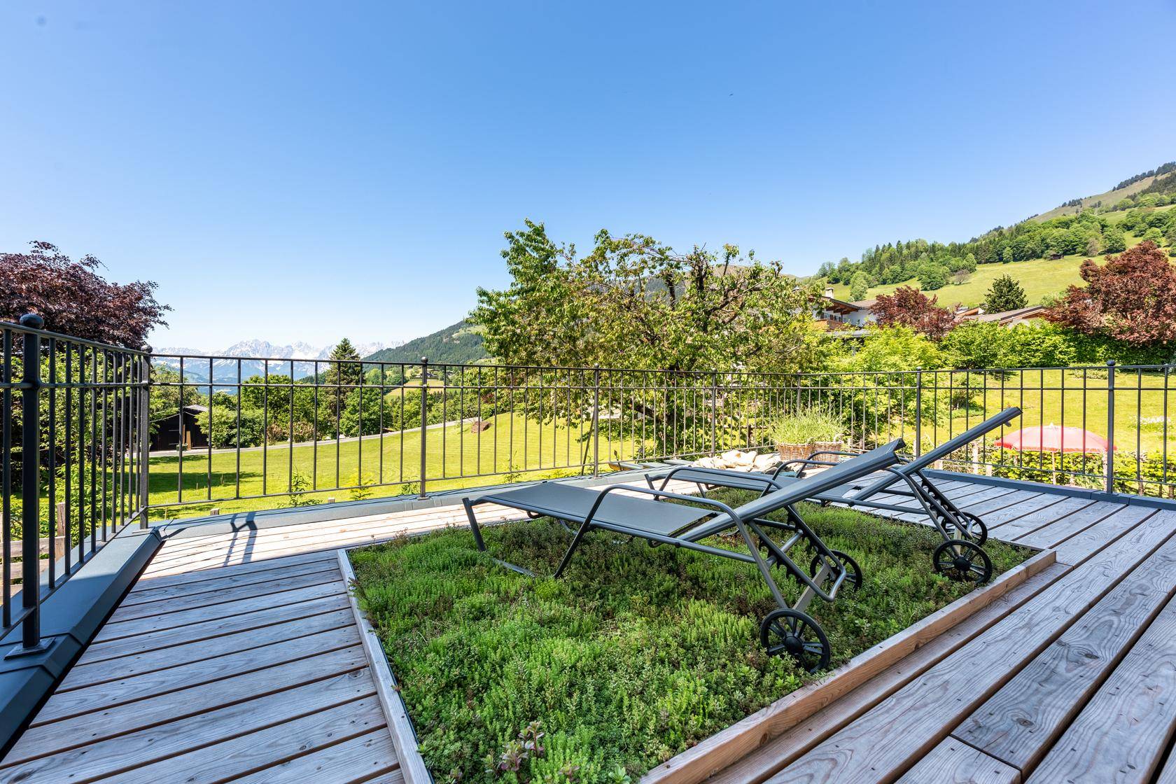 Sonnige Holzterrasse mit Liegestühlen und begrünten Beeten, umgeben von grüner Landschaft.