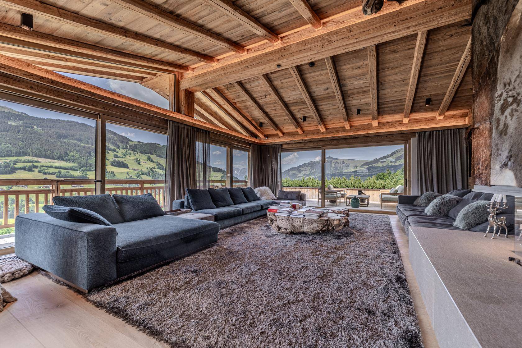 Großzügiges Wohnzimmer mit Panoramablick auf die Berge, Holzbalkendecke und gemütlichem Teppichboden.