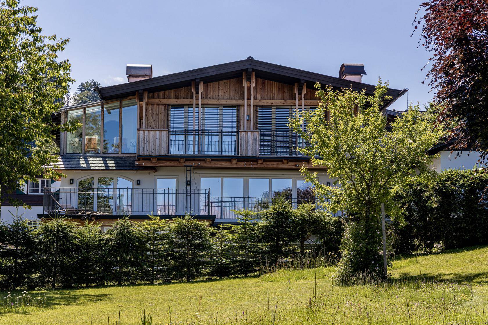 Moderne Villa mit alpinen Holzelementen, großen Fenstern und Balkonen, umgeben von Grünflächen.