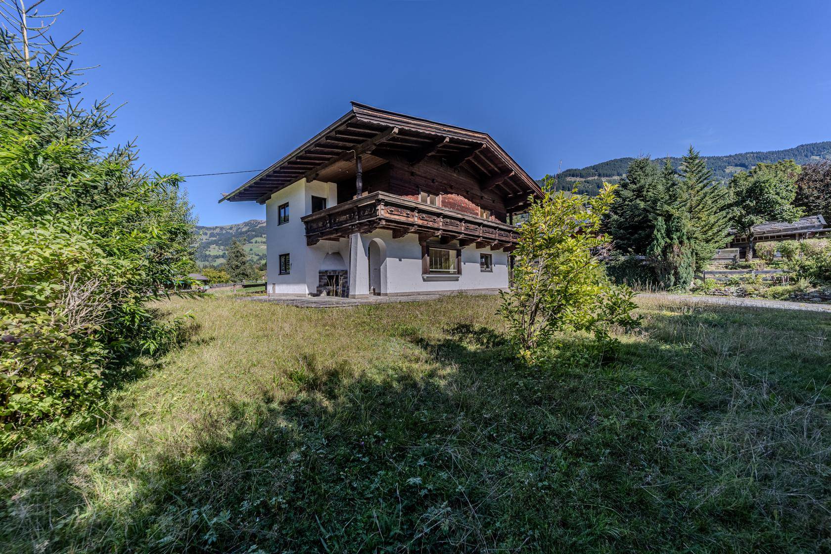 Traditionelles Haus im alpinen Stil mit Holzbalkon, umgeben von grünen Wiesen und Bergen.