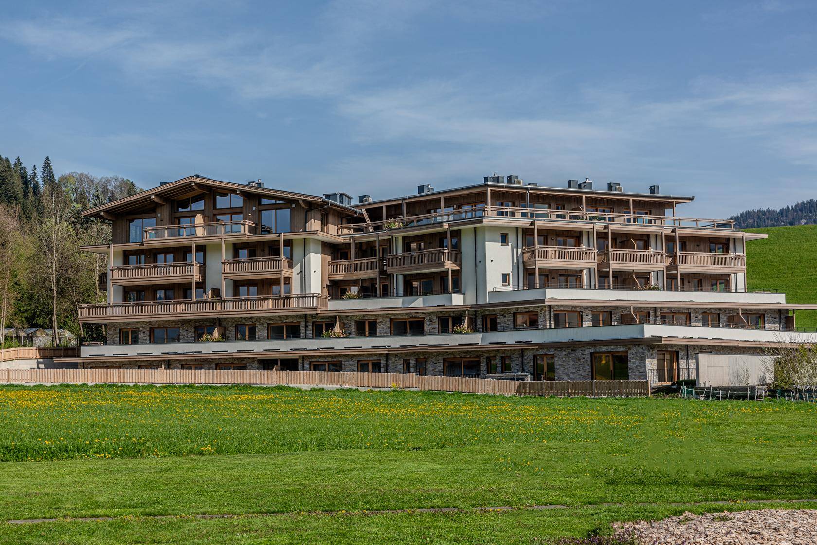 Großes, modernes Apartmentgebäude mit Holzfassade und zahlreichen Balkonen, umgeben von grünen Wiesen.