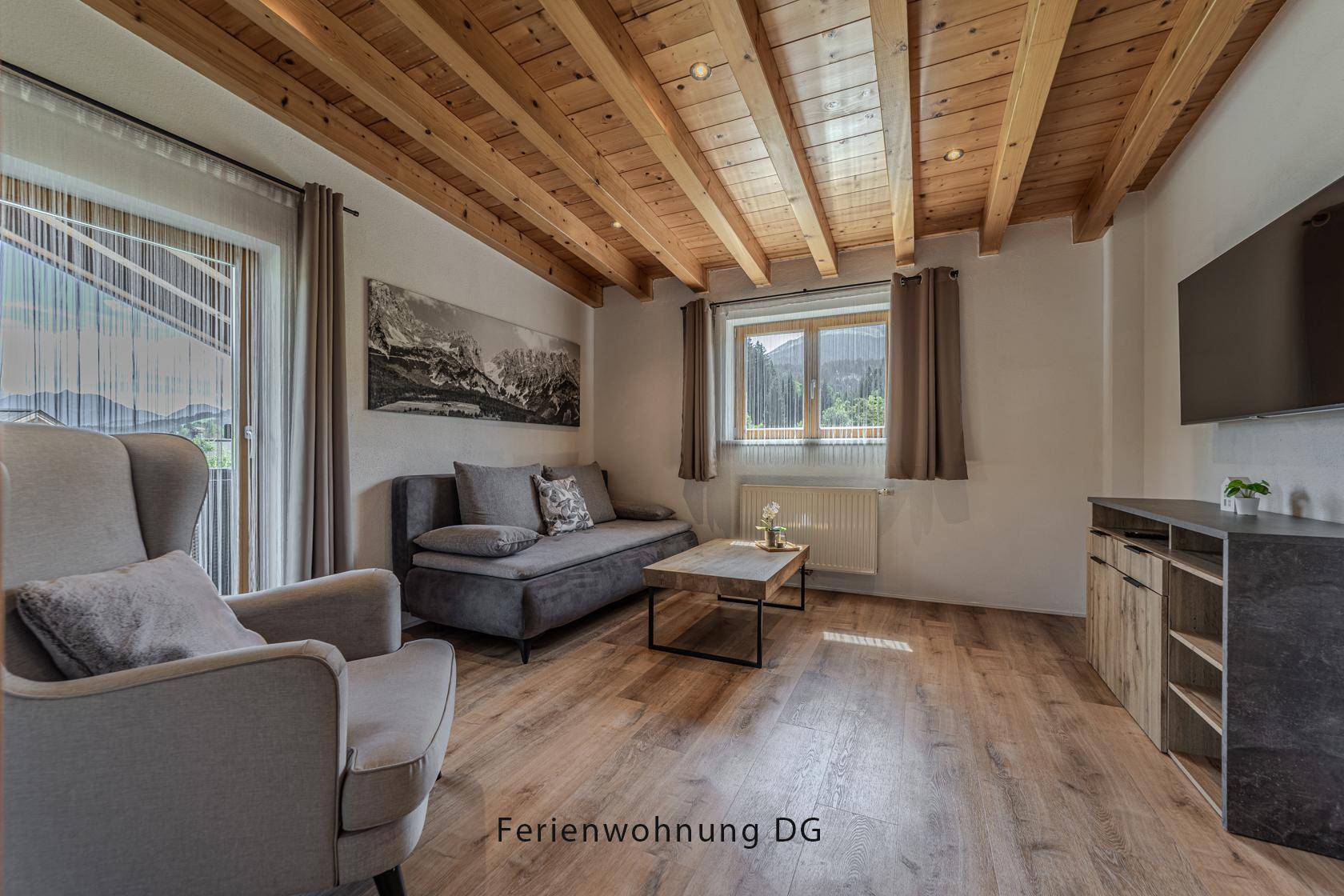 Gemütliches Wohnzimmer mit Holzbalkendecke, Sofa und Fernseher, hell und einladend.