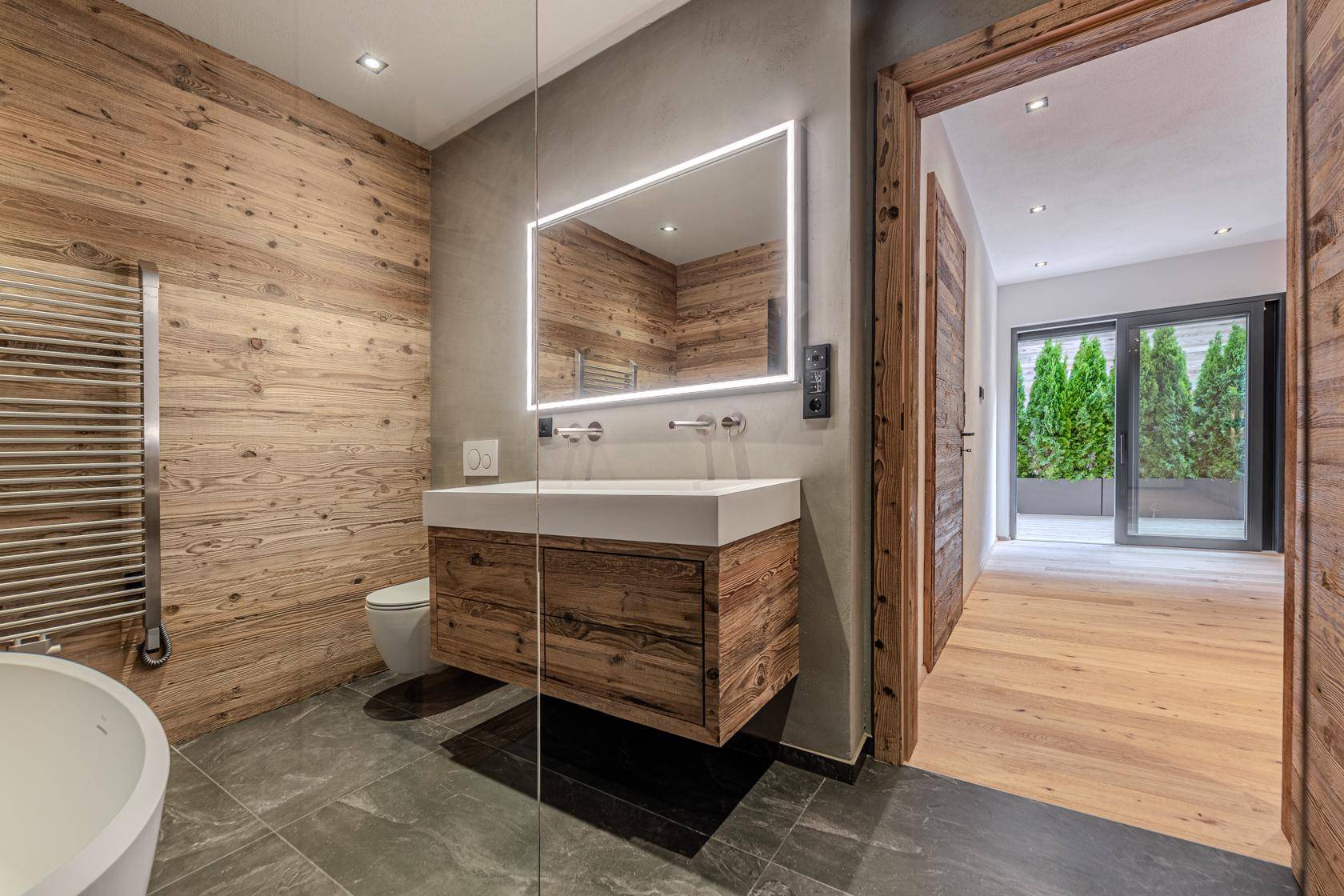 Modernes Badezimmer mit beleuchtetem Spiegel, Holzwaschtisch und Zugang zum Schlafzimmer.