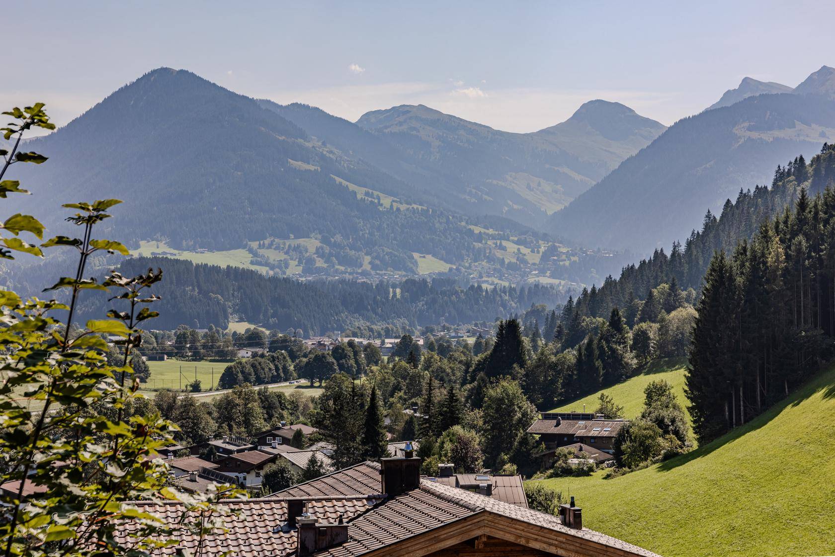 Weitläufiger Ausblick über ein idyllisches Tal mit bewaldeten Bergen, Wiesen und traditionellen Häusern.