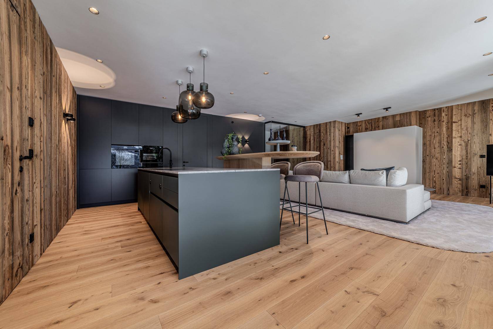 Moderne Küche mit dunklen Fronten und offener Verbindung zum Wohnbereich mit Holzwänden.