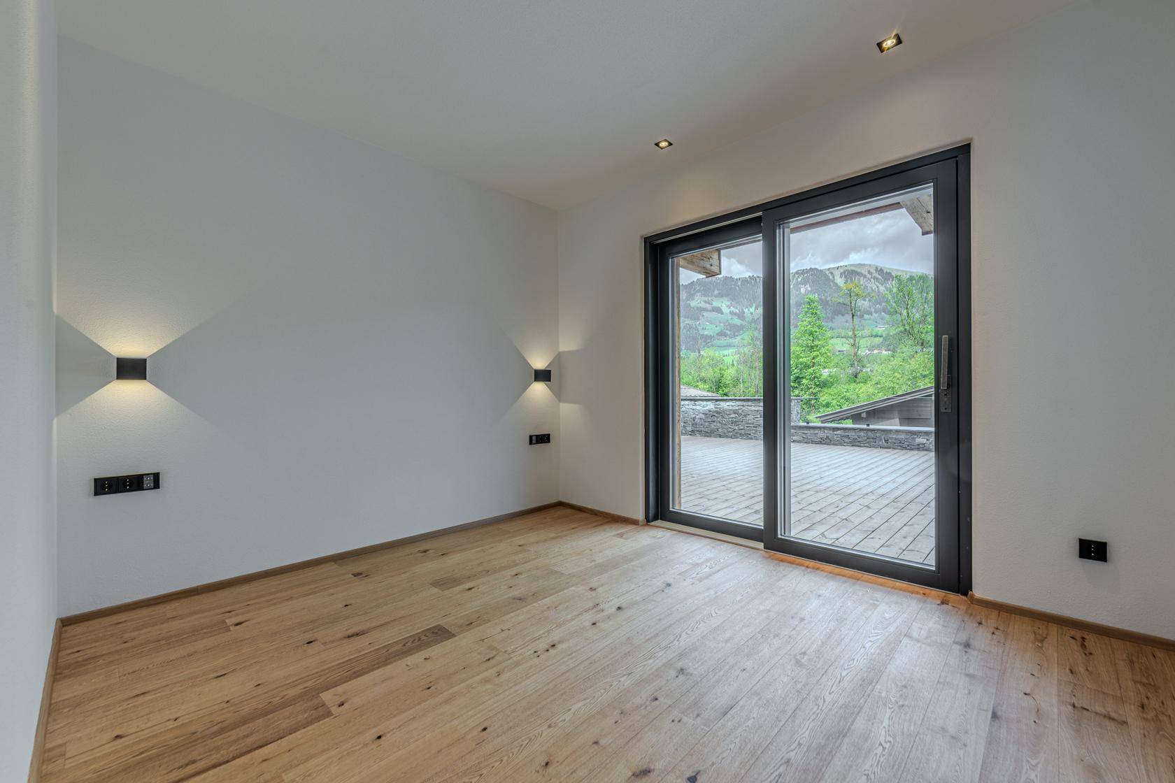 Helles Schlafzimmer mit Holzboden und großem Schiebefenster zur Terrasse mit Bergblick.