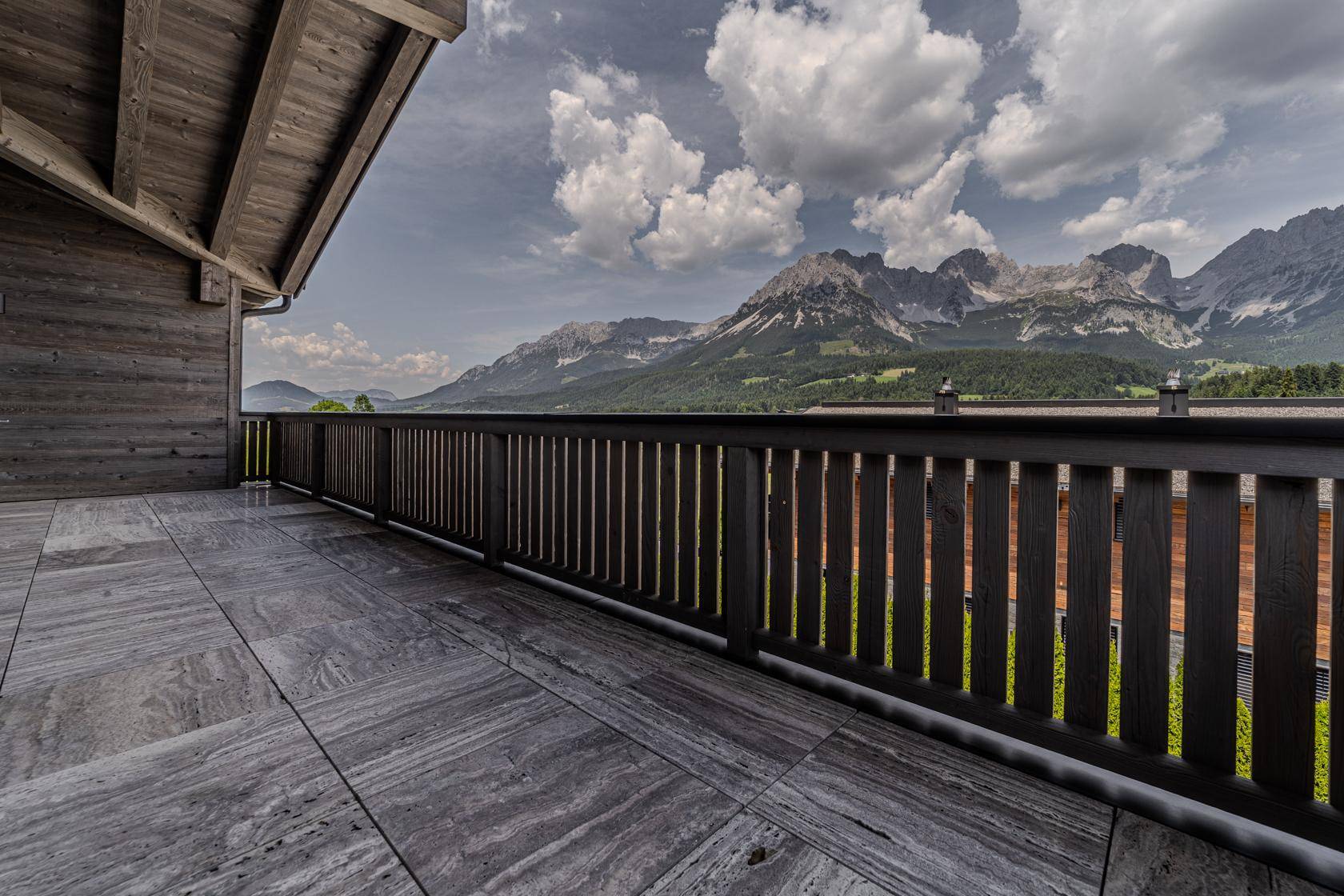 Großer Balkon mit Steinfliesen und Holzgeländer, bietet einen atemberaubenden Panoramablick auf die Berge.
