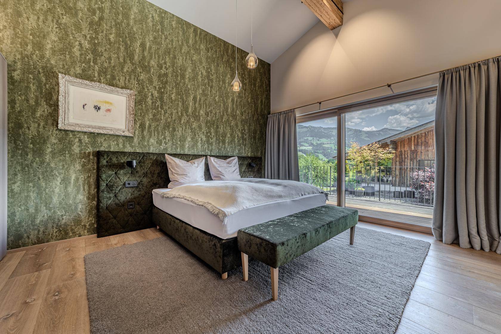 Schlafzimmer mit grüner Akzentwand, Holzboden und großen Fenstern mit Zugang zum Balkon und Bergblick.