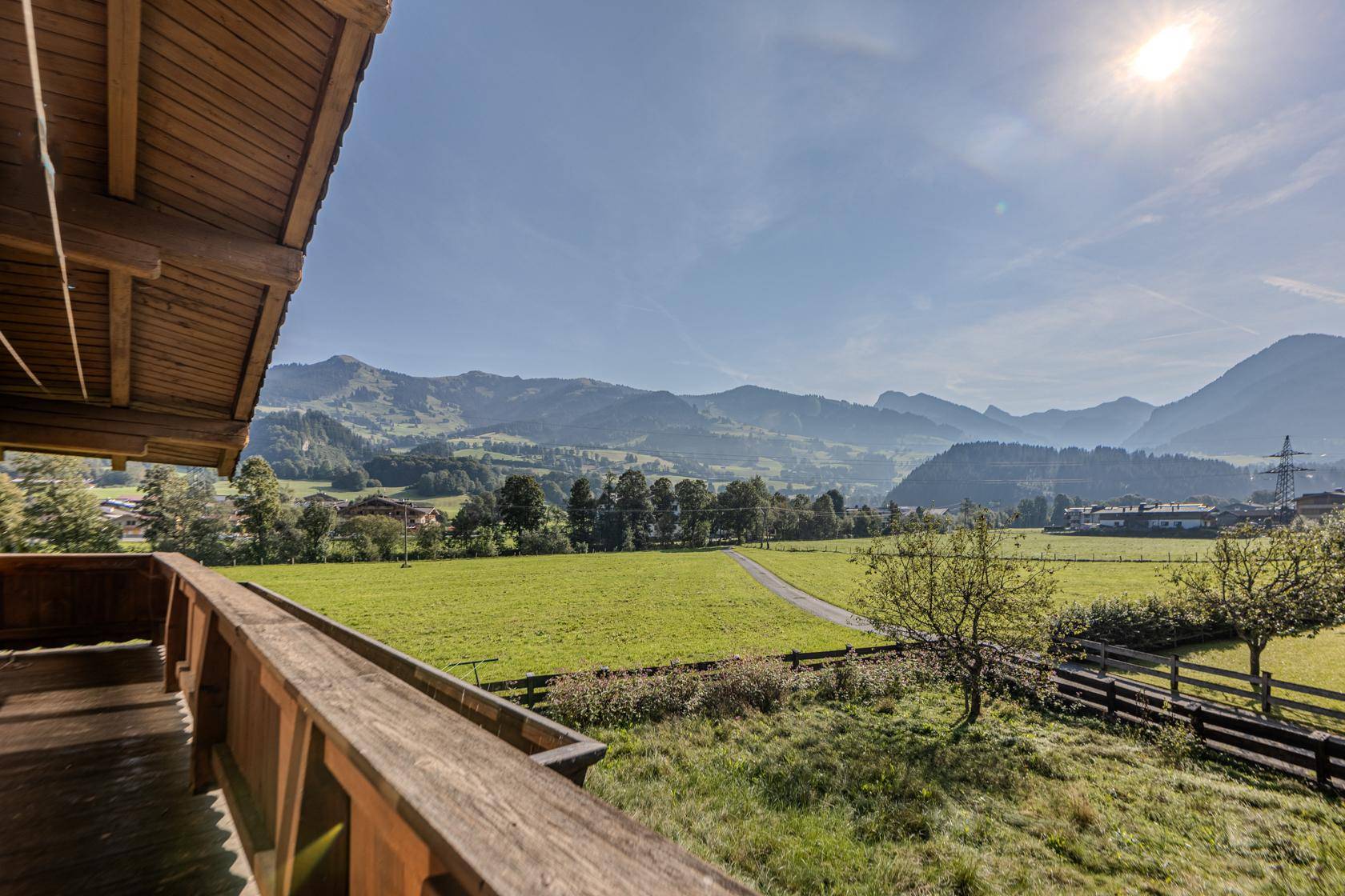 Sonniger Balkon mit Holzboden und weitem Blick über grüne Wiesen und Berge.
