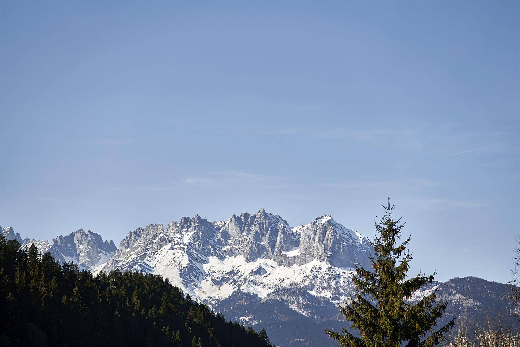 Majestätische Berglandschaft mit schneebedeckten Gipfeln und grünen Wäldern unter blauem Himmel.