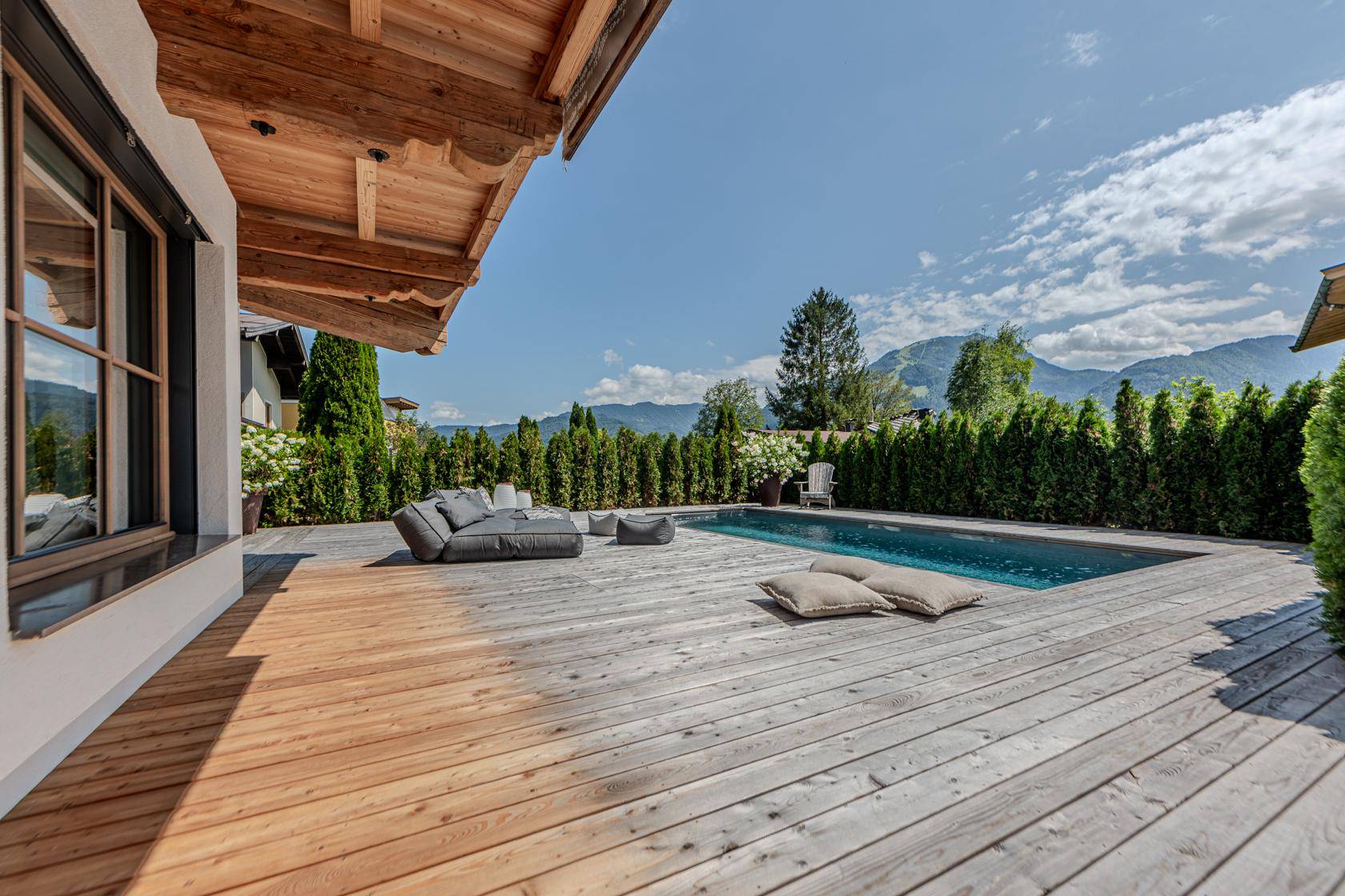 Geräumige Holzterrasse mit Pool und bequemen Lounge-Möbeln, umgeben von einer grünen Hecke und Bergblick.